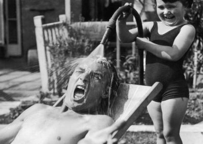 Uma menina sente grande prazer ao molhar seu pai com água fria da mangueira do jardim enquanto ele tomava sol em um dia quente. 1936.