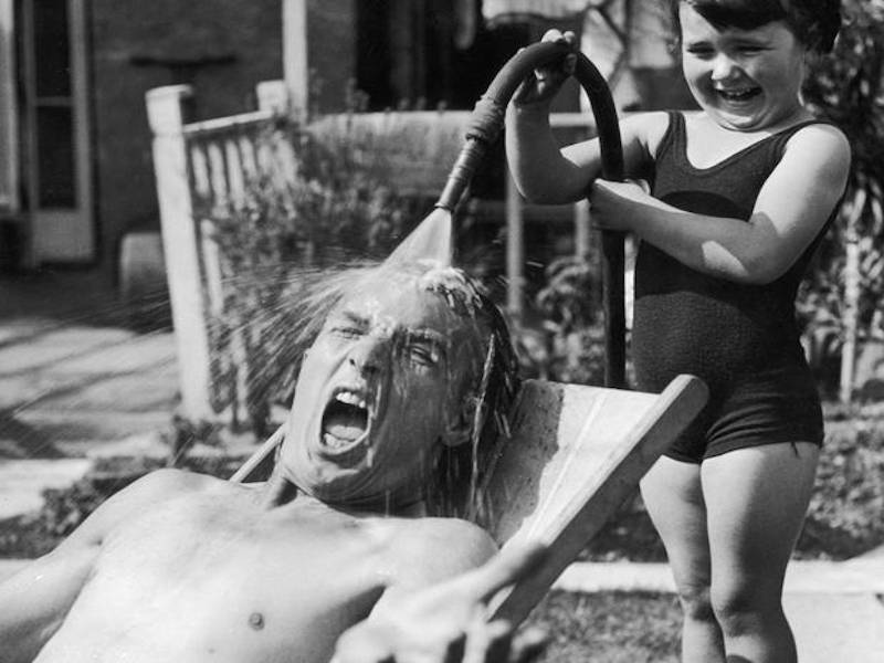 Uma menina sente grande prazer ao molhar seu pai com água fria da mangueira do jardim enquanto ele tomava sol em um dia quente. 1936.